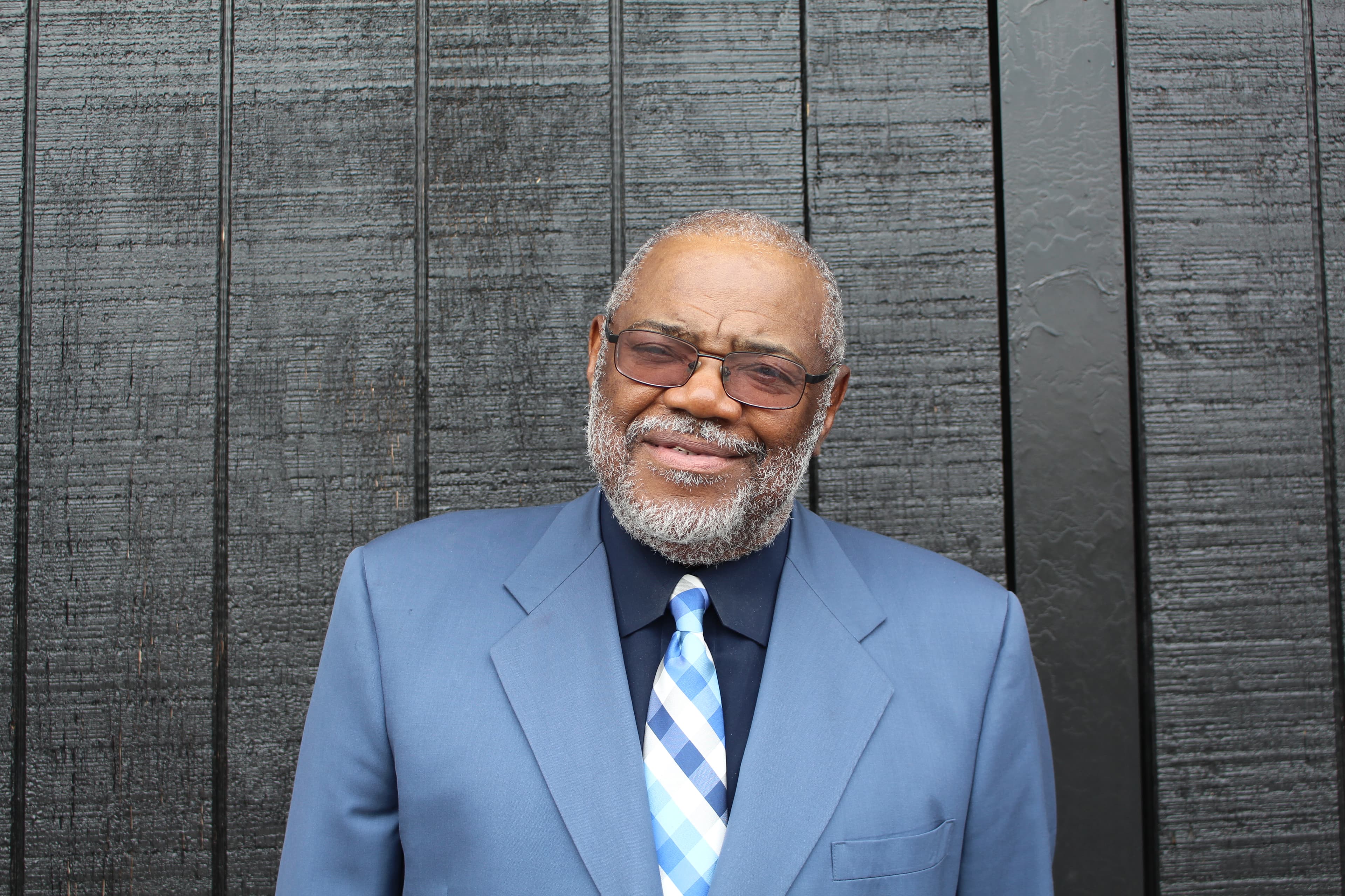 Smiling older Black man with a grey beard wearing a blue suit and checkered tie.