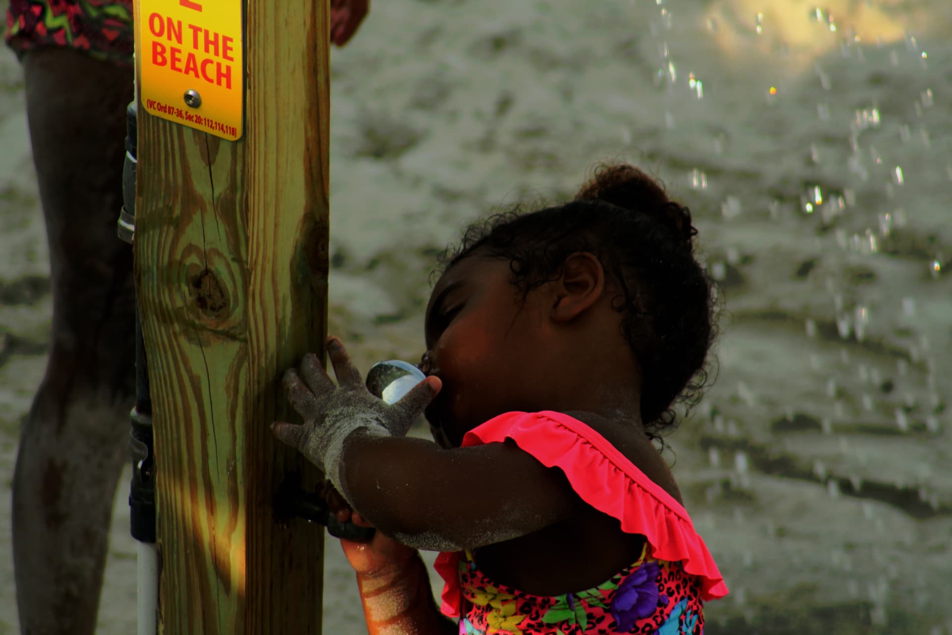Young girl in a pink swimsuit drinks from a water fountain on a sandy beach.