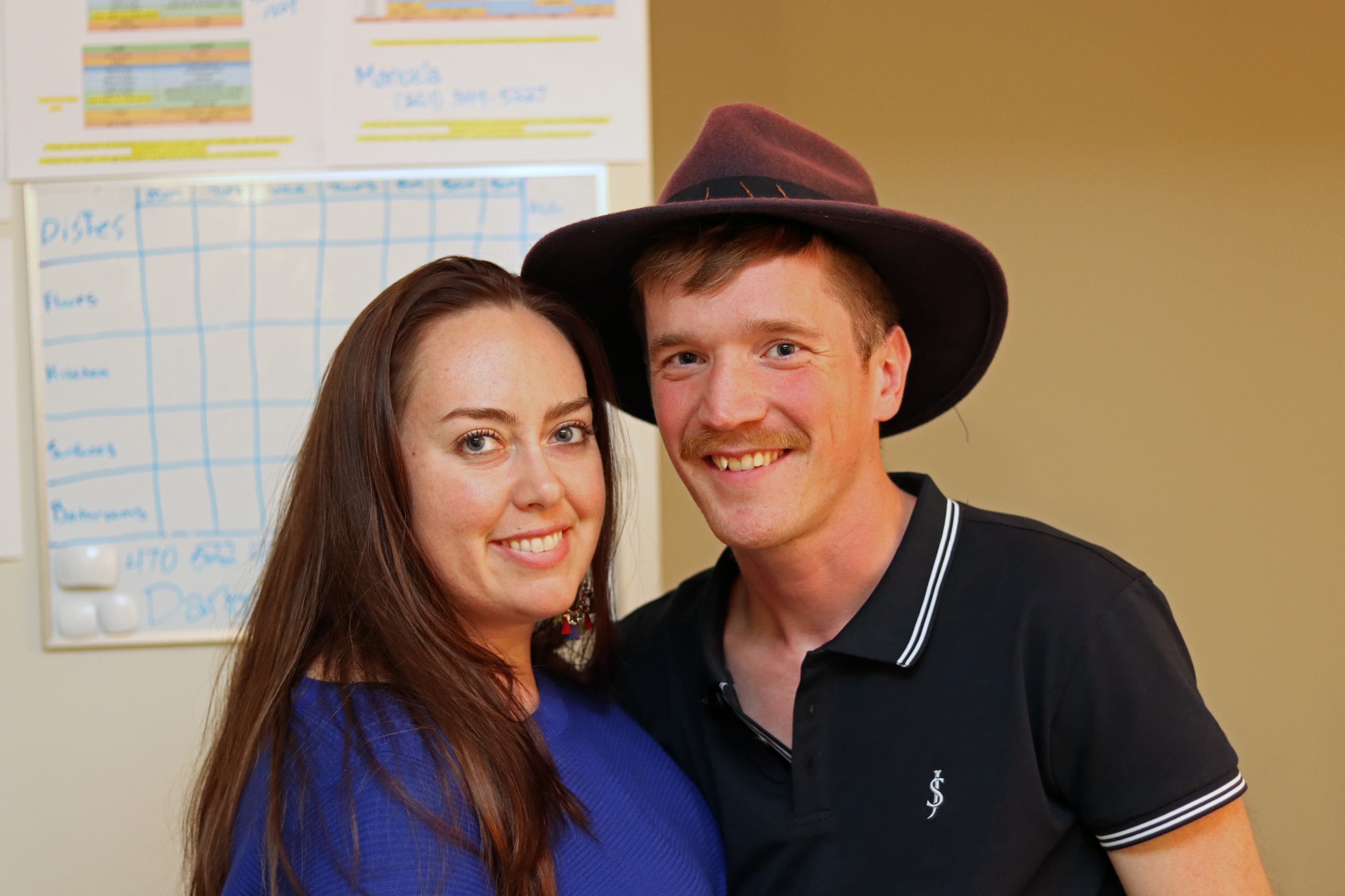 Smiling woman in blue and a man with a mustache and brown hat posing together.